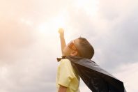 A child wearing sunglasses and a black cape raises one arm towards the sky, set against a cloudy background. The scene conveys a sense of adventure and imagination, potentially relating to themes like the present perfect Spanish.