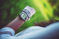 A close-up of a wrist adorned with multiple silver bracelets, featuring a large round watch with a light-colored dial, set against a blurred green background.