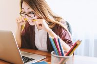 A woman with long hair and glasses biting a pencil while looking at a laptop, sitting at a desk with colored pencils in a holder and a notebook.