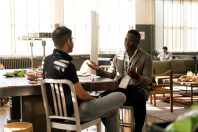 Two people engaged in a conversation at a table in a bright, modern space. One person is seated in a light sweater, gesturing while speaking, and the other is listening attentively, wearing a dark shirt. A variety of refreshments are visible on the table.