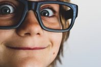 Close-up of a smiling child with short blonde hair wearing large black glasses. The child's wide eyes and cheerful expression are prominent, set against a light background.