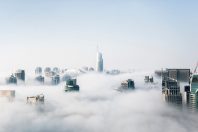 A panoramic view of a city skyline partially shrouded in fog, with tall buildings emerging from the clouds. The foreground features various architectural styles, emphasizing the modern urban environment.