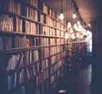 A cozy interior view of a library with tall shelves filled with books. Warm pendant lights hang along the ceiling, illuminating the space and creating a serene reading atmosphere.