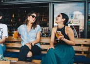 Two women sitting on a wooden bench at a bar, laughing and enjoying drinks. One woman wears sunglasses and a denim shirt, while the other wears a black top and a green skirt, both appear to be in a lively atmosphere.