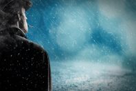 A man wearing a dark coat and hat stands outdoors in the snow, looking away from the camera. Snowflakes cascade down against a blurred blue background, creating a cold, wintry atmosphere. The scene may represent winter weather in Spanish.