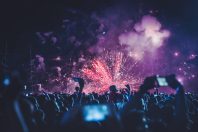 A crowd of people enjoying a fireworks display at night, with colorful sparks bursting in the sky. The atmosphere is festive, with several individuals capturing the scene on their smartphones. This relates to celebratory events and holidays in Spanish.