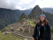 A woman smiling in front of Machu Picchu, with lush green mountains in the background and cloudy skies above. The ancient ruins of Machu Picchu are visible, highlighting the site's historical significance.