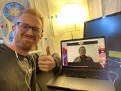A man with glasses giving a thumbs-up while sitting at a desk with a laptop showing a smiling person on a video call. The workspace features a lamp and a fan in the background. This scene represents learning Spanish through online interaction.