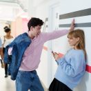 A young man in a pink hoodie leans against a wall with one hand while a young woman in a blue sweater stands next to him, looking at her phone. The background shows a hallway with people walking.