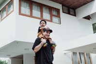 A man carrying a child on his shoulders outside a modern house. The child is smiling and wearing a black beanie, while the man is dressed in a black t-shirt and has colorful shoes. The house has large windows and a minimalist design.