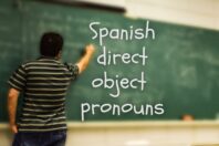 A teacher writing on a green chalkboard, with the words 'Spanish direct object pronouns' written in large white letters.