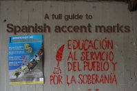A wall displaying a painted sign that reads "A full guide to Spanish accent marks." Below, there is text in red stating "EDUCACIÓN AL SERVICIO DEL PUEBLO Y POR LA SOBERANÍA" alongside a poster for a seminar on political economy.
