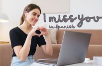A woman smiling while making a heart shape with her hands during a video call. A laptop is open in front of her, with text overlay saying "I miss you in Spanish".