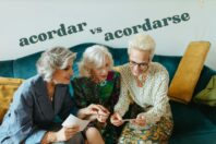 Three older women sitting on a green sofa, examining photographs together. The words 'acordar vs acordarse' are displayed above them, emphasizing a topic related to expressions of courtesy in Spanish. Each woman is wearing stylish clothing.