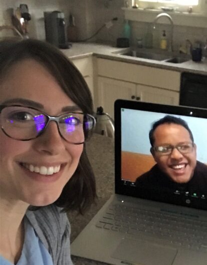 A woman with glasses smiling at a laptop showing a smiling man on a video call. The setting is a kitchen with countertops and a sink in the background.