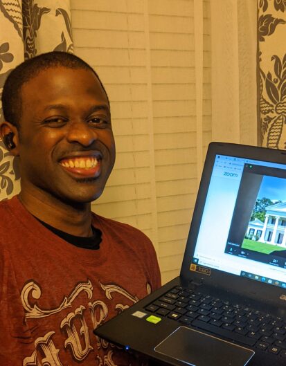 A man smiling while holding a laptop displaying a Zoom video call with an image of a large house on the screen. The setting includes light-colored window blinds in the background.