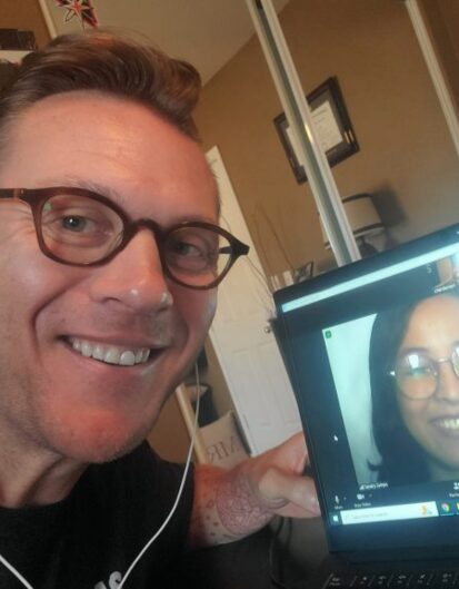 A man with glasses smiling while taking a selfie, with his laptop showing a woman in glasses smiling back during a video call. The background includes a door and a glimpse of room decor.