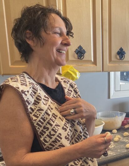 A woman with short curly hair wearing a patterned apron smiles while standing in a kitchen. There are various cooking ingredients and utensils visible on the counter behind her.