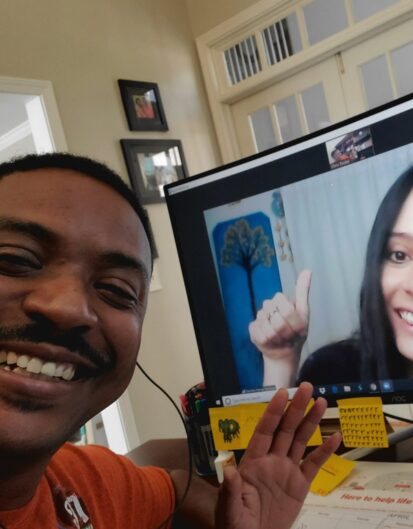 A man smiling and posing for a selfie while video chatting with a woman on a computer screen. The background shows a bright room with decorations and a desk with various items.