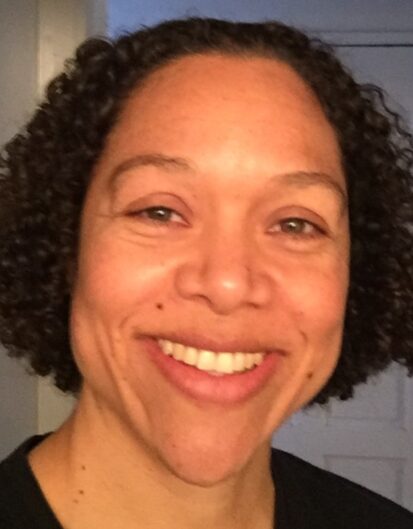 A close-up portrait of a woman with curly hair, smiling while looking directly at the camera. She has a natural appearance and is wearing a black shirt.