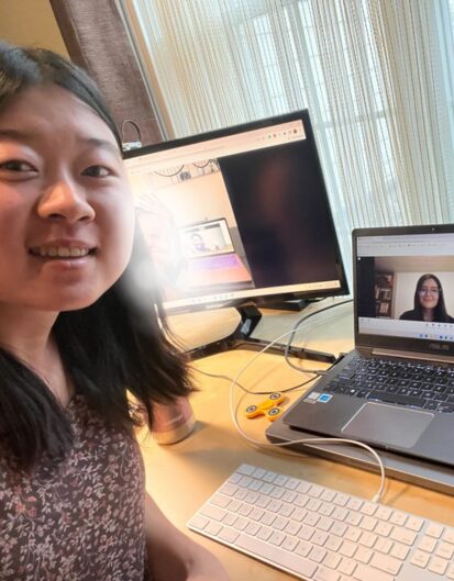 A young woman smiling at the camera while participating in a video call, sitting at a desk with a laptop and an external monitor displaying another person on the screen. A keyboard is visible in front.