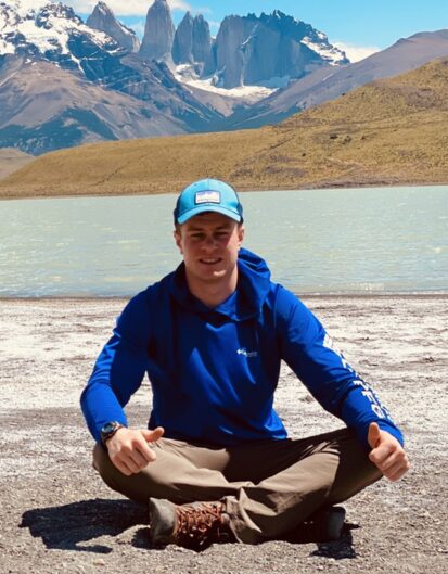 A person sitting cross-legged on the shore of a lake, wearing a blue long-sleeve shirt and a blue cap. Behind, dramatic mountains rise under a clear blue sky with patches of snow.