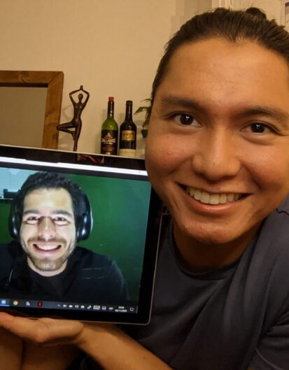 A person smiling while holding a tablet that displays another person smiling on a video call. In the background, there are bottles on a shelf and a statue figure.