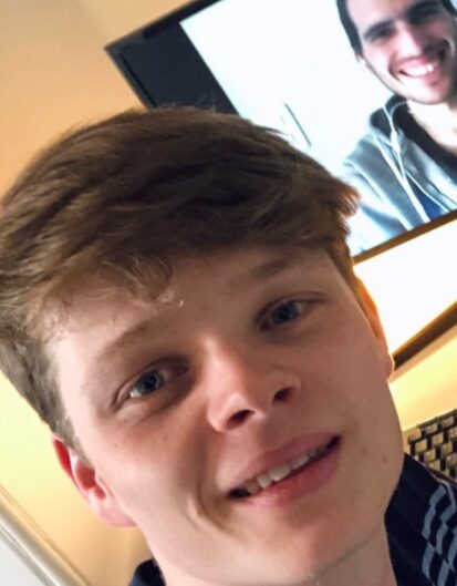 A young man smiling at the camera with short, light brown hair. In the background, a person is smiling on a computer screen.
