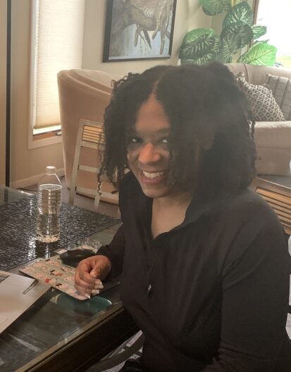 A woman with curly hair sitting at a table, smiling while looking at the camera. A water bottle and papers are on the table in a well-lit room with indoor plants in the background.