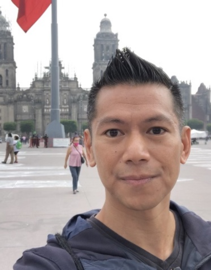 A man with short, styled hair takes a selfie in a plaza with historic buildings in the background. A large flag is visible to the left, and people walk around the square.