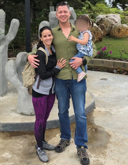 A man and a woman posing together outdoors with a young child in a floral dress. The man is wearing a green shirt and jeans, while the woman is dressed in a black and gray outfit. They stand in front of a decorative stone structure.