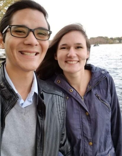 Two smiling individuals standing close together near a body of water. One person is wearing glasses and a leather jacket, while the other is wearing a purple coat. The background shows trees and water.