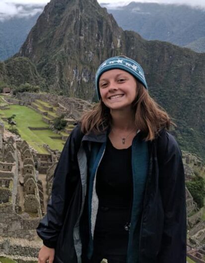 A young woman smiling while standing in front of the ancient ruins of Machu Picchu, surrounded by lush green mountains under a cloudy sky. She is wearing a blue beanie and a black jacket.