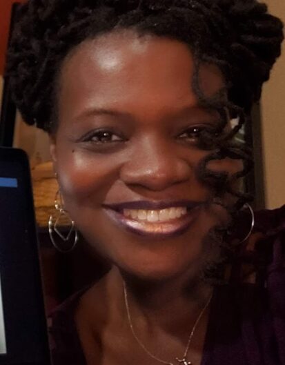 A woman with curly hair and hoop earrings smiles at the camera while holding a laptop. The background shows a softly lit room with warm tones.