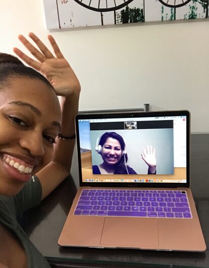 A woman smiling and waving at a laptop screen, which shows another woman wearing headphones and waving back during a video call.