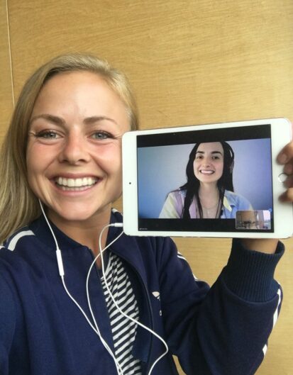 A woman with long hair and a sporty outfit smiling as she holds a tablet displaying another woman smiling during a video call. Both are in well-lit environments.