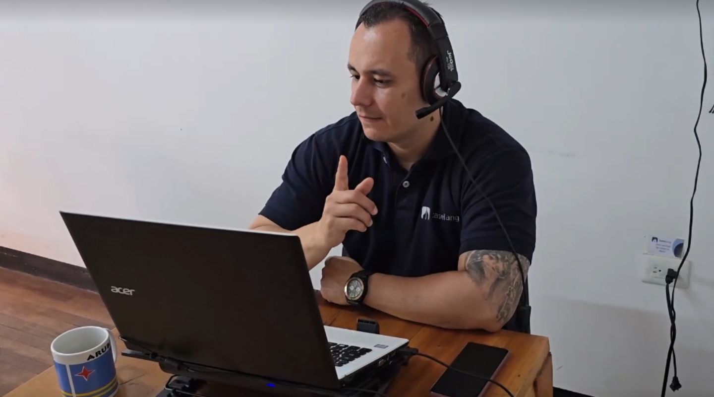 A man wearing a headset is sitting at a wooden desk, gesturing with one hand while looking at a laptop. A coffee mug and a smartphone are also on the desk.