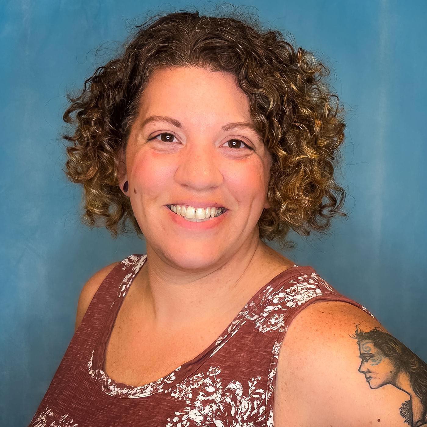 A woman with curly hair wearing a maroon floral top smiles against a blue background. She has a tattoo on her right shoulder. The image conveys a cheerful and friendly demeanor.