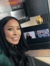 A woman with long black hair smiles at the camera while sitting at a desk with a laptop displaying a video call. The background features a lamp and framed posters on the wall.