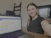 A woman in a black shirt takes a selfie at a desk with an open laptop, a large TV, and a shelving unit in the background.